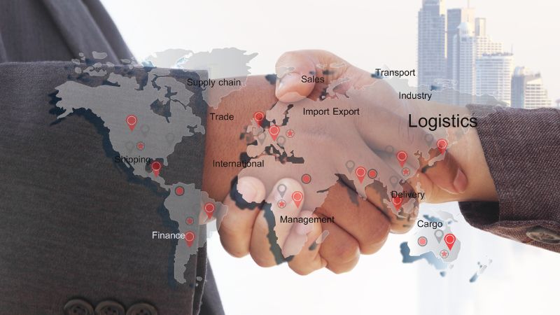 Two people shake hands over a map with logistics, trade terms, and red pins showing global connections, with city buildings in the background.