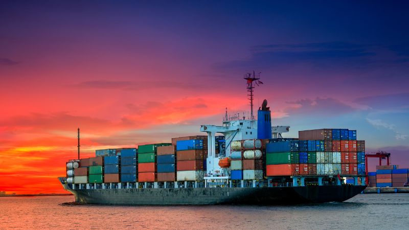 A cargo ship with containers crosses calm water at sunset, under a colorful sky.