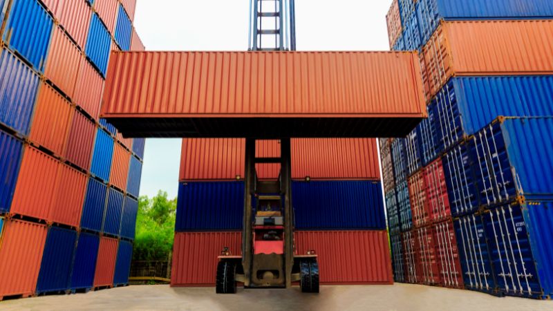 A forklift lifts a red container among stacks of red and blue containers in an outdoor storage area.