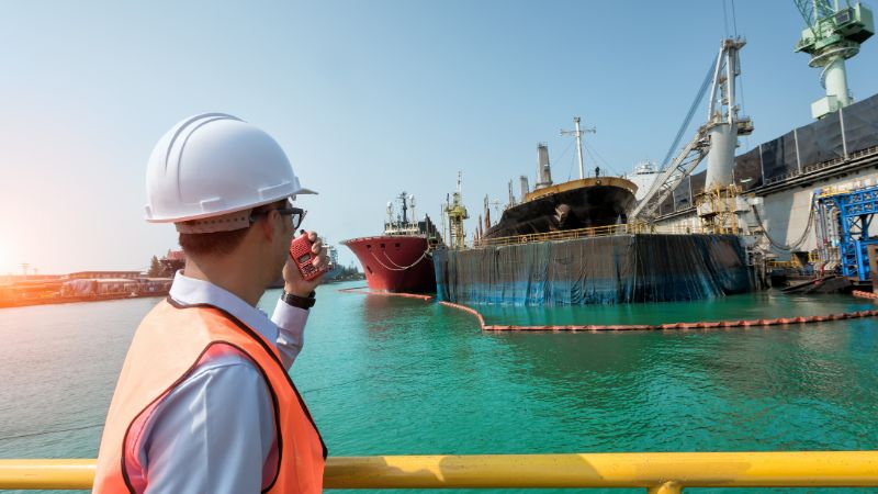 A worker in a hard hat and vest communicates via radio, overseeing a docked ship partially surrounded by barriers at an industrial harbor.